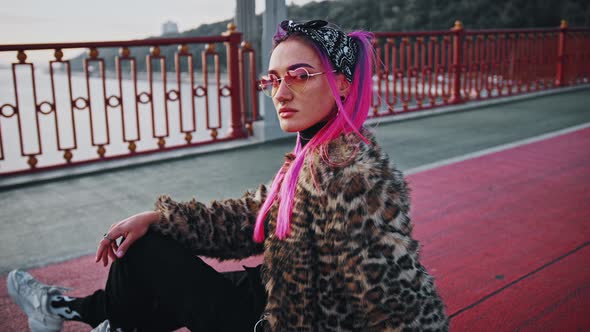 Young Hipster Female with Pink Hair Wearing Informal Outfit is Sitting Sideways on Asphalt While