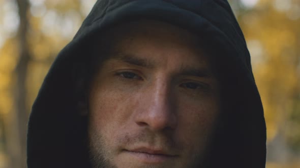 Close Up Portrait of Young Serious Bearded Guy Wearing Hood Looking at Camera at Autumn Park