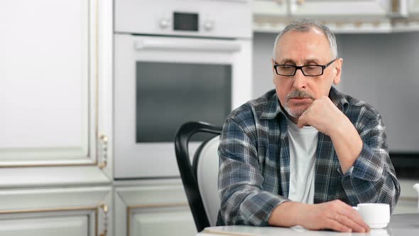 Portrait of Pensive Grayhaired Casual Elderly Man Sitting at White Kitchen During Coffee Break