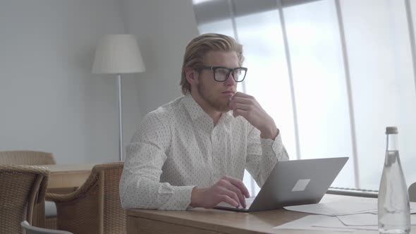 Blond Thoughtful Man in Glasses Sitting at the Table in a Light Comfortable Office in Front