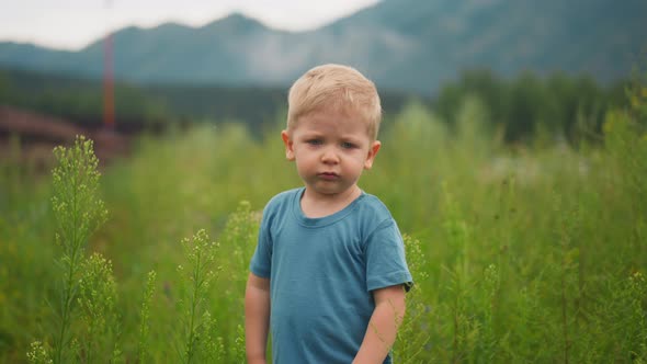 Upset Little Boy Stands Among High Green Grass in Lush Field