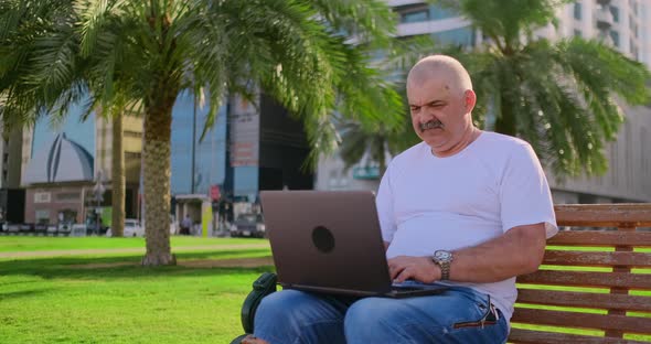 Senior Man Sitting in Park with Laptop in Summer and Chatting