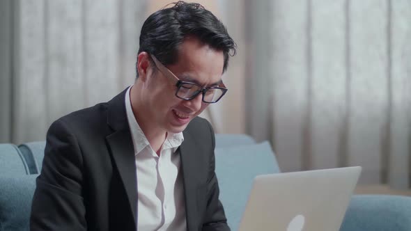 Close Up Of Smiling Asian Businessman In Jacket And Shorts Sitting On Sofa And Typing On A Laptop