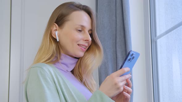 Cheerful blonde woman using mobile phone and listening to favorite music at home in 4k