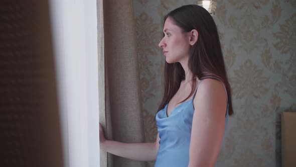 Young Woman Stands Near the Window in the Bedroom and Enjoys the Sunrise