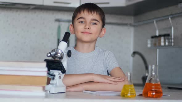 Portrait of a Smart Schoolboy Who Sits at a Table at Home Next To a Microscope and Flasks with