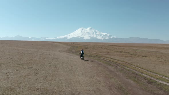 Woman Tourist Rides Mtb Bicycle Along Empty Rural Road in Field Against Snowy Elbrus Mountain Under