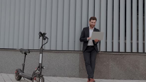 Smiling Modern Businessman in Formal Wear Stands with Laptop and Work Near Escooter