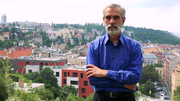 Old Senior Man Looks To Camera with Serious Face - City (Buildings) in Background