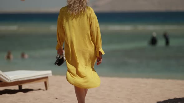 a Curly Blonde in a Yellow Fluttering Dress Walks Along a Sandy Beach to the Sea and Carries Sandals