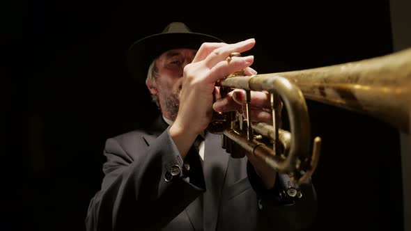 Man in hat and jacket plays trumpet on dark background, front view. Musician plays trumpet in studio