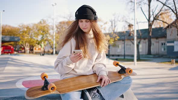 Young Stylish Female Student with a Skateboard in Her Hands Sits Outside and Listens to Music From