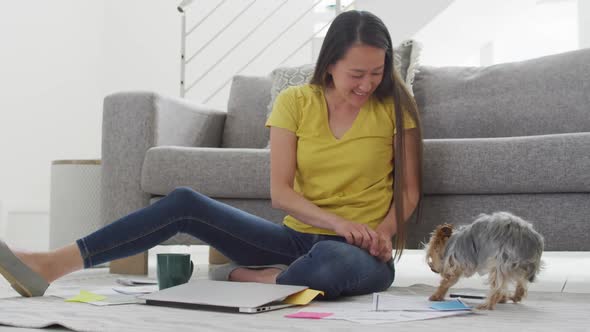 Happy asian woman sitting on floor, working remotely from home with laptop, petting dog