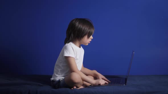 Boy Child in a White Tshirt and Shorts is Sitting on a Bed Against a Blue Wall of the House