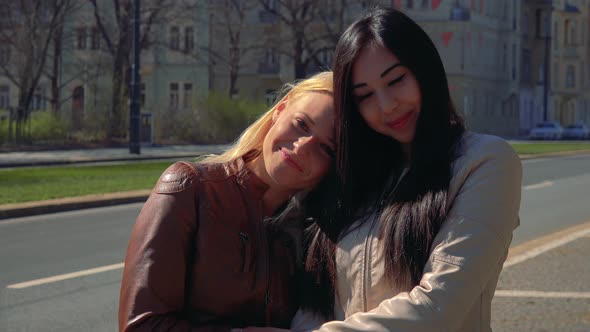 A Young Caucasian Woman and a Young Asian Woman Hug and Smile at the Camera in a Street