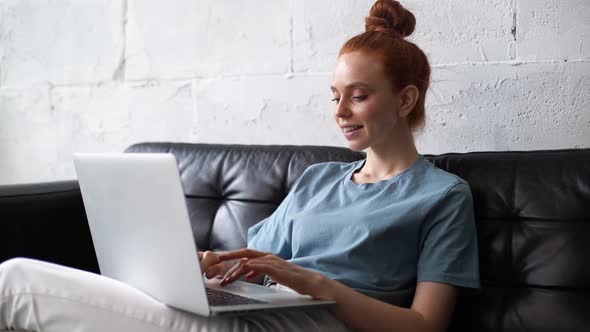 Cheerful Redhead Young Businesswoman Is Working on Modern Laptop Computer While Sitting on Couch.