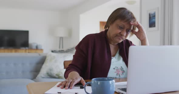 Worried african american senior woman at dining table, using laptop and holding head