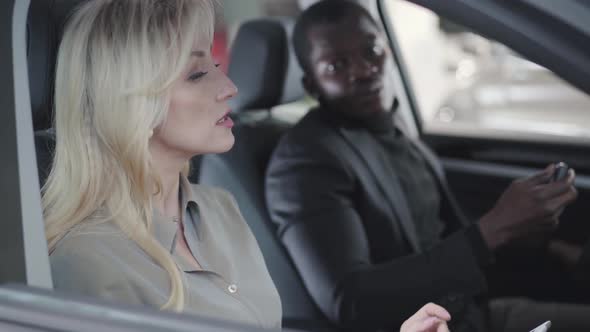 Side View of Beautiful Blond Caucasian Woman Sitting in Car Salon with Tablet and Explaining