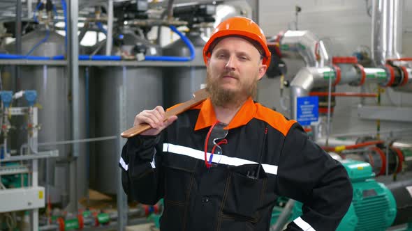 A Bearded Industrial Mechanic in an Orange Hard Hat Stands at a Workplace in a Production Workshop
