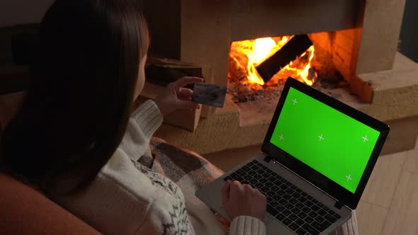 Woman Holds a Credit Card While Using a Green Screen Laptop To Place an Order Near the Fireplace