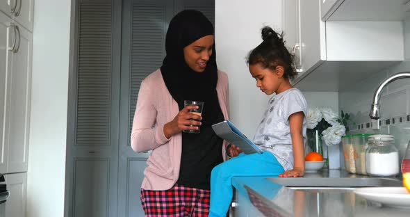 Young mother with a hijab drinking a glass of water near her daughter in the kitchen 4k