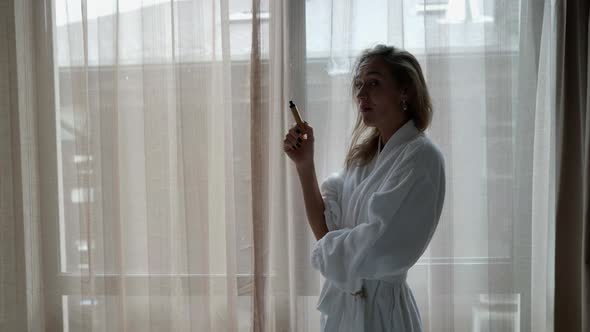 a Woman in a White Coat Stands at a Large Window with Light Curtains and Smokes an Electronic Cigar