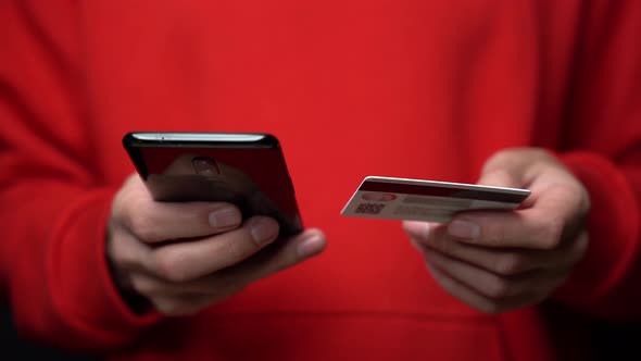 Close-up of a man in a red hoodie holding a smartphone and a credit card in his hand