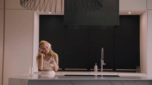 A Young Woman Dancing with a Phone in Her Hands and Headphones in the Kitchen