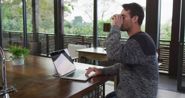 Caucasian man working at home, using laptop and drinking coffee in dining room