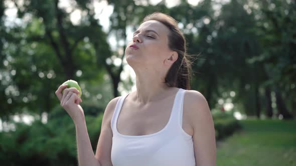 Portrait of Beautiful Joyful Sportswoman Eating Healthful Green Apple and Smiling at Camera. Happy