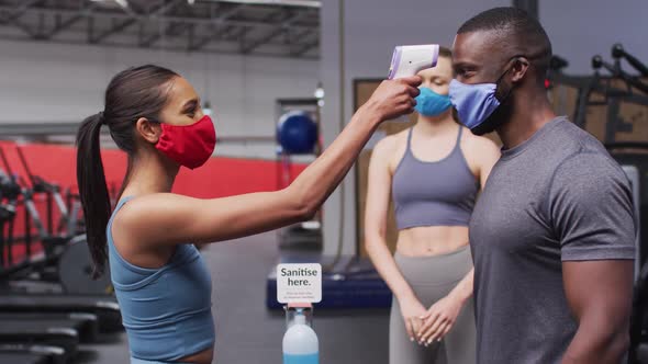 Fit caucasian woman wearing face mask measuring temperature of fit african american man in the gym