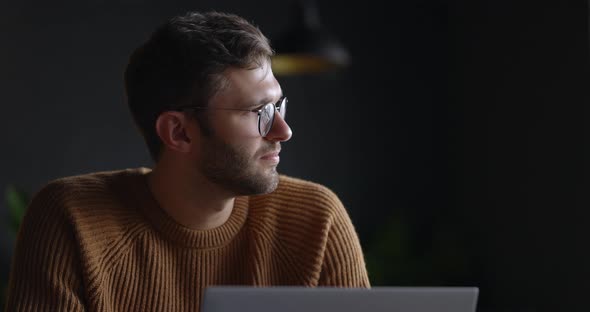 A Closeup View of Thoughtful Serious Man in Eyeglass Sit with Laptop Thinking of Solution at Dark