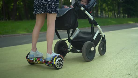 A Young Mother Carries a Stroller While Standing on a Hoverboard in the Park