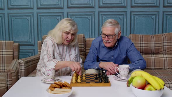 Retired Senior Couple Talking Drinking Tea Playing Chess in Modern Living Home Room Lounge Together
