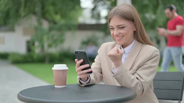 Young Businesswoman Celebrating Success on Smartphone in Outdoor Cafe