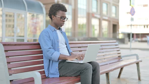 Young African Man Celebrating Success on Laptop While Sitting Outdoor on Bench