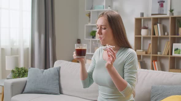Cheerful Woman Eats a Donut at Home in the Living Room and Drinks Coffee