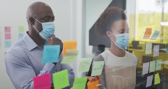 Diverse businessman and businesswoman in face masks brainstorming by board with memo notes in office