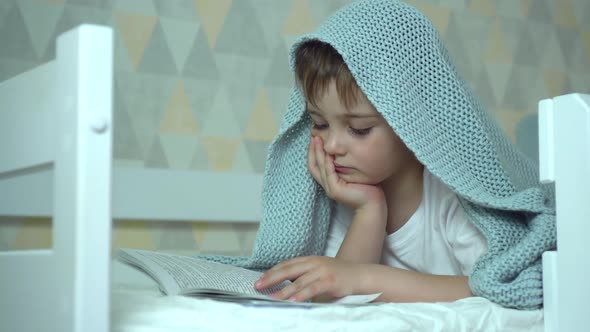 a Small Boy Reads a Book Lying Under a Blanket