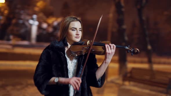 A gorgeous girl in a bright dress and black coat performs classical music on a violin in a winter