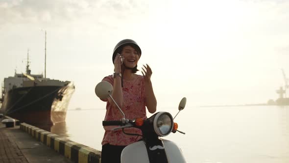 Young Beautiful Girl Speaking on Phone Near Sea on Scooter at Sunrise