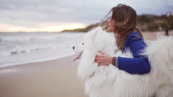 Side View of a Young Woman Sitting on the Sand and Embracing Her Dog of the Samoyed Breed By the Sea