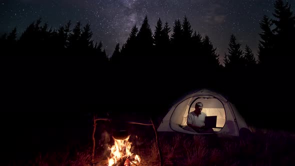 Man Works Behind a Laptop in a Tent Near a Fire at Night