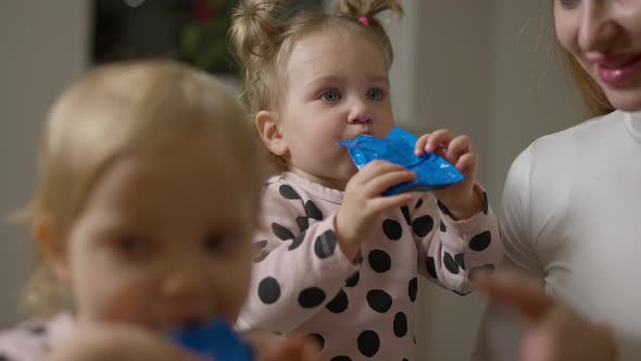 Cute Baby Girl Drinking Multivitamin Juice with Twin Sister and Mother at Home