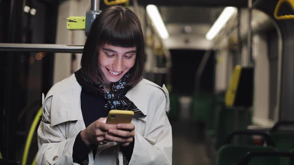 Portrait of Happy Young Woman Using Smartphone Standing in Public Transport. City Lights Background.