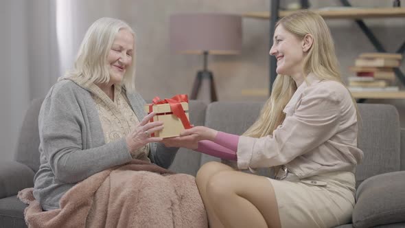 Cheerful Senior Woman Giving Gift Box to Older Friend Sitting on Couch in Living Room