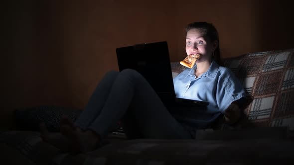 girl works at the laptop late at home and eats pizza on a sofa