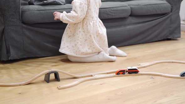 Toddler Girl in White Dress Plays with Wooden Train at Home in the Living Room