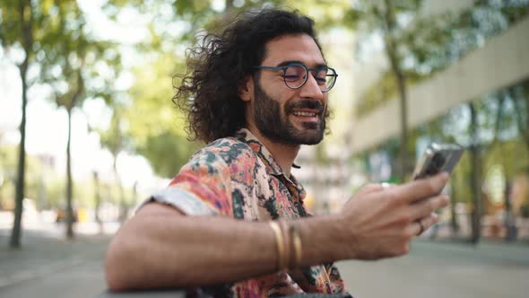 Pleased curly-haired bearded man in eyeglasses typing by mobile