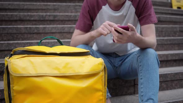 Unrecognizable Young Man Sitting on Stairs with Yellow Food Delivery Backpack Surfing Internet on